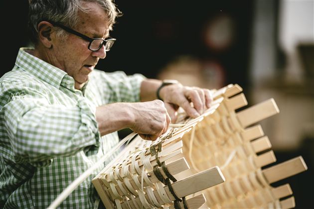 Ein älterer Mann mit Brille arbeitet an einem Holzstuhl. Er ist konzentriert und verwendet verschiedene Techniken zur Fertigung.