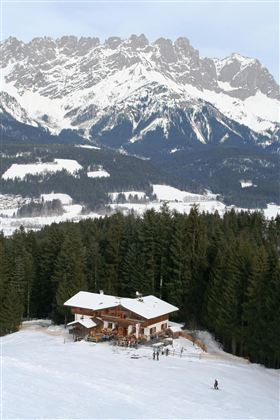 A cozy mountain cabin in the snow surrounded by fir trees and majestic mountains. The sky is cloudy and the landscape appears peaceful.
