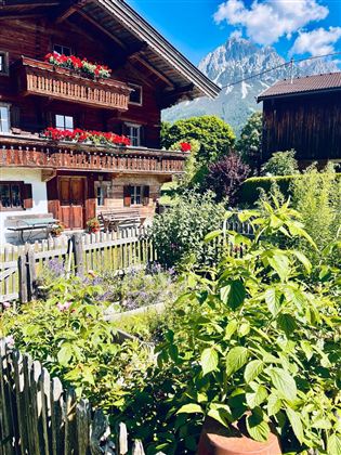 Ein traditionelles Chalet mit blühenden Balkonblumen und einem liebevoll gepflegten Garten. Im Hintergrund sind majestätische Berge und ein blauer Himmel zu sehen.