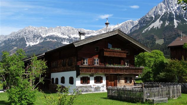 A traditional farmhouse in the mountains with snow-covered peaks in the background. The surroundings are green and inviting, ideal for rural recreation.