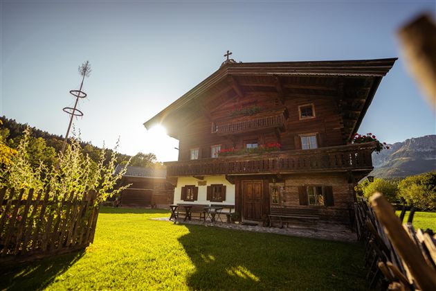 Eine gemütliche Stube mit Holzwänden und zwei Fenstern. Ein runder Tisch mit einer weißen Tischdecke steht in der Mitte, umgeben von Bankauflagen.