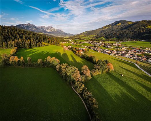 Zwei Wanderer gehen auf einem Pfad durch eine grüne Wiese. Im Hintergrund sind Bäume und eine Hütte, beleuchtet von der Sonne.