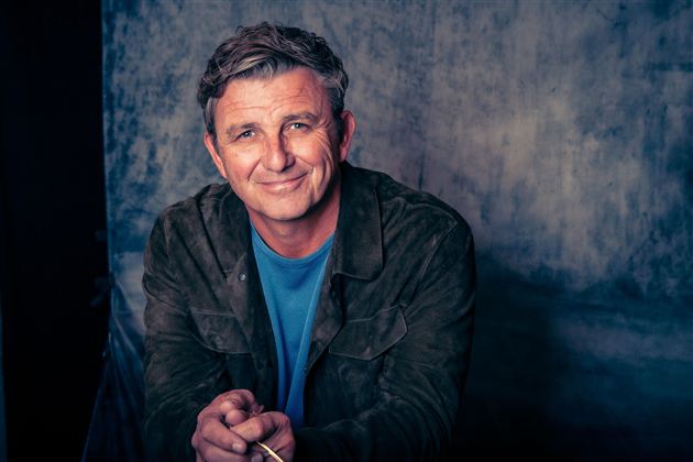 Ein lächelnder Mann mit grau melierten Haaren sitzt vor einem neutralen Hintergrund. Er trägt eine Jacke und ein T-Shirt und schaut freundlich in die Kamera.