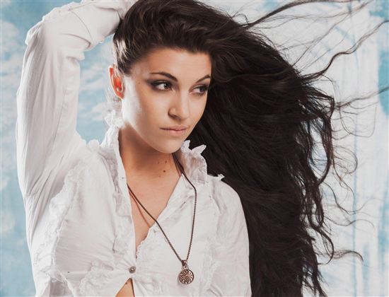 Eine junge Frau mit langen, fließenden Haaren trägt ein weißes Hemd. Sie blickt nachdenklich und hat eine Kette um den Hals.