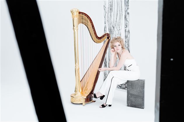 A woman sits next to a golden harp in a minimalist, bright room. She is wearing an elegant white outfit and appears thoughtful.