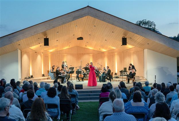 Eine Musikerin in einem roten Kleid steht im Mittelpunkt einer Bühne und singt. Das Publikum sitzt gespannt davor, umgeben von einer schönen Abendkulisse.
