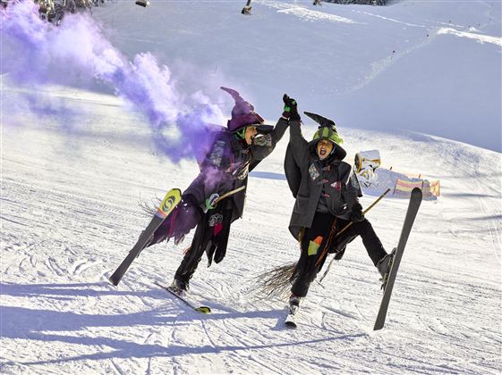 Twee verklede heksen vieren op de skipiste en houden elkaars handen vast. Kleurige rookfakkels omringen hen in het besneeuwde landschap.
