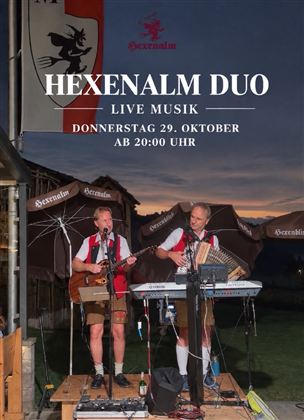 Two musicians are performing live on an outdoor stage. In the background, a beautiful sunset and the mountains can be seen.