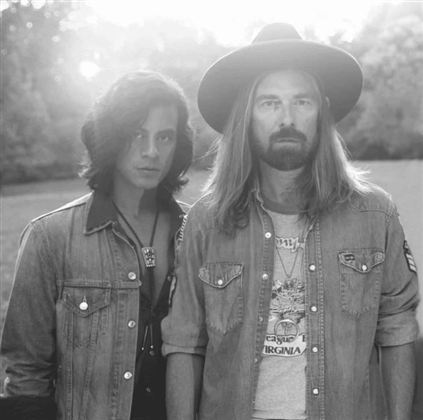 Two men are standing close together and looking at the camera. They are wearing casual clothing and have stylish haircuts in a natural, sunlit background.