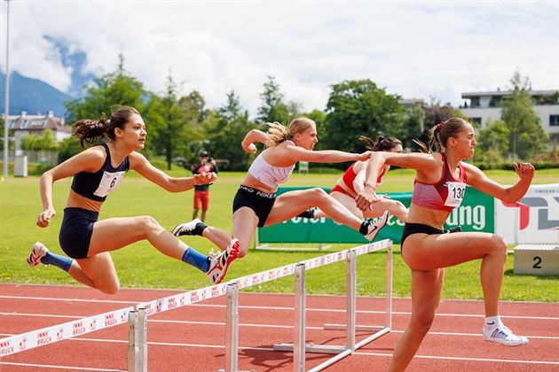 Eine Gruppe von Frauen springt über Hürden auf einer Laufbahn. Der Hintergrund zeigt eine grüne Landschaft unter einem klaren Himmel.