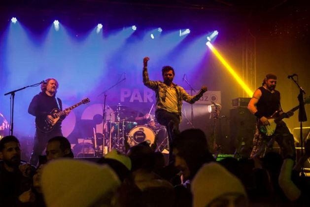 A band plays live on stage while the crowd dances and cheers. The lights are colorful and create an energetic atmosphere.