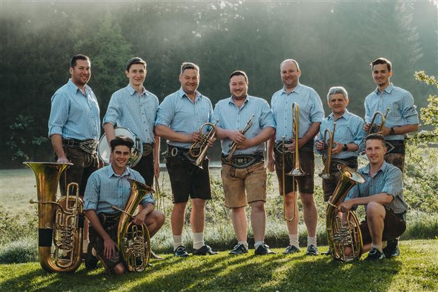 Een groep muzikanten staat in de open lucht en draagt traditionele klederdracht. Ze houden verschillende blaasinstrumenten vast en glimlachen naar de camera.