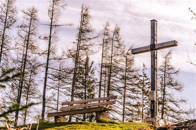 A wooden bench stands next to a wooden cross, surrounded by bare trees. The sky is slightly overcast.