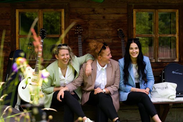Drei Frauen sitzen lächelnd auf einer Bank vor einem Holzhaus. Im Hintergrund sind Gitarren und Pflanzen zu sehen.