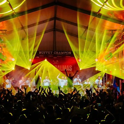 Eine große Menschenmenge genießt ein Konzert unter einem Zelt. Bunte Lichter und Strahlen schaffen eine festliche Atmosphäre.