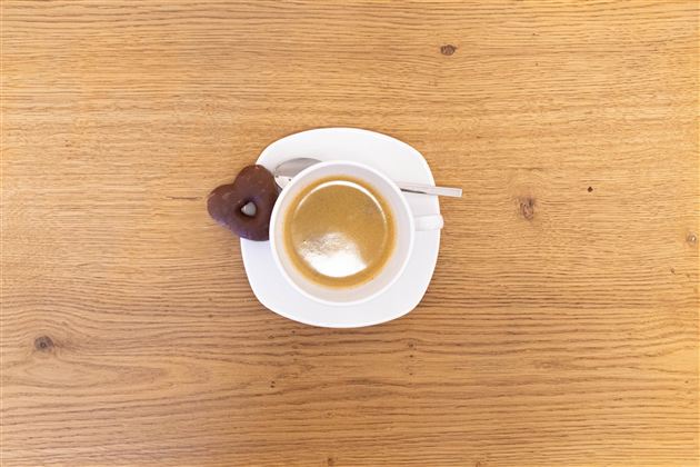 Šálek kávy na bílém talíři s malým srdíčkovým sušenkou. Stůl je z světlého dřeva.