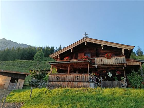 Ein traditionelles Holzhaus in den Bergen, umgeben von grünen Wiesen. Vor dem Balkon sind bunte Blumen und eine Kreuzdekoration zu sehen.