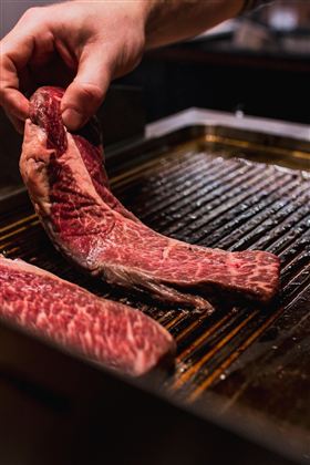 Ein Koch hebt ein Stück rotes Fleisch auf einen Grill. Die Fleischstücke zeigen eine schöne Marmorierung.