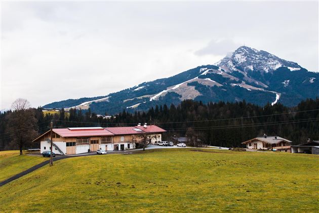 Ein malerisches Landschaftsbild mit einem Bauernhaus im Vordergrund und einem majestätischen Berg im Hintergrund. Der Himmel ist bewölkt und die Wiesen sind grün.