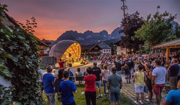A picturesque village view at night with mountains in the background. People gather in the square under bright lights.