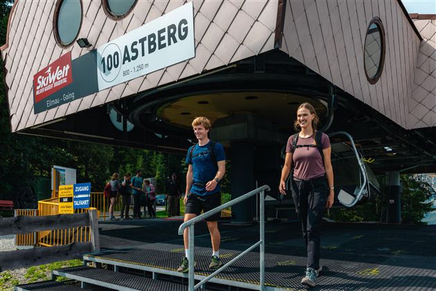 Zwei Wanderer verlassen eine moderne Seilbahnstation. Im Hintergrund sind weitere Personen zu sehen, die sich auf den Weg machen.