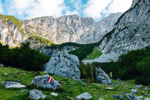 Eine beeindruckende Berglandschaft mit hohen Felsen und einer grünen Wiese. Am Hang sind Wanderzeichen sichtbar.