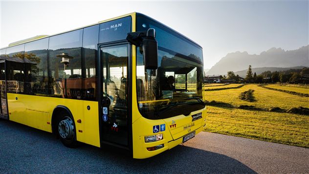 Ein gelber Bus steht auf einer Straße neben einem Feld. Im Hintergrund sind Berge und ein klarer Himmel zu sehen.