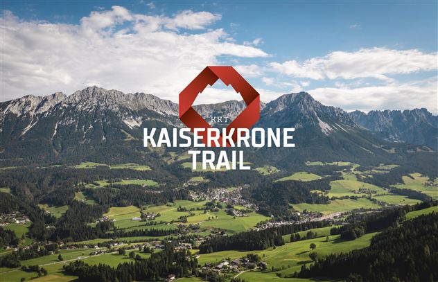 An impressive mountain landscape with green meadows and high mountains in the background. In the foreground stands the logo for the Kaiserkronen-Trail.