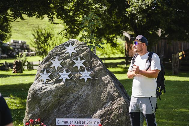 Ein Mann steht neben einem großen Fels mit Sternen. Im Hintergrund sind Bäume und eine Wiese zu sehen.