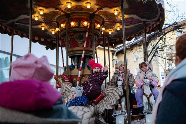 Een draaimolen met blije kinderen die op kleurrijke paarden zitten. Op de achtergrond zijn volwassenen te zien die het gebeuren observeren.