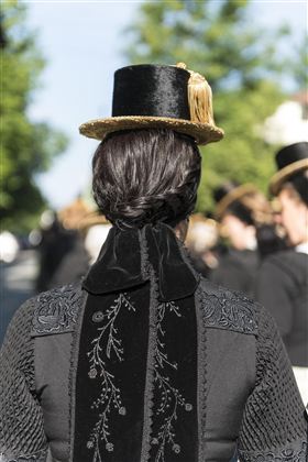 Eine Person mit einem traditionellen schwarzen Kleid und einem eleganten Hut steht im Vordergrund. Im Hintergrund sind weitere Personen in ähnlicher Kleidung zu sehen.