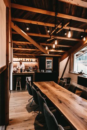 Ein modernes Esszimmer mit einem großen Holztisch und stilvollen Stühlen. Im Hintergrund ist eine offene Küche mit einem Koch zu sehen.