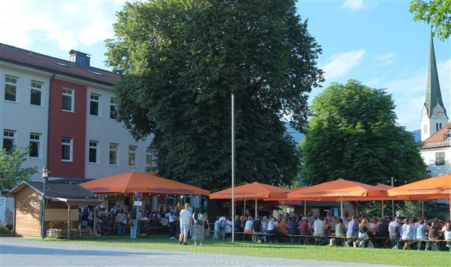 A bustling outdoor dining area with orange umbrellas and many guests. In the background, a large building and a church tower are visible.