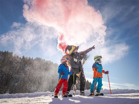 Een vrolijke wintersportdag met kinderen die op een besneeuwde helling staan. Iemand in een kostuum wijst met een gekleurde rookgranaat naar de lucht.