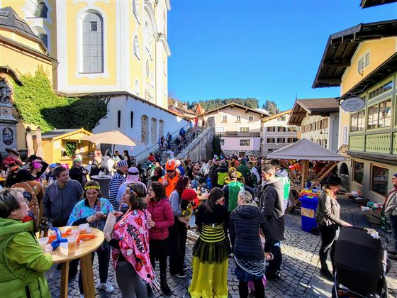 A lively crowd is celebrating in a picturesque old town. Many are dressed in colorful costumes, surrounded by historic buildings and a festive atmosphere.