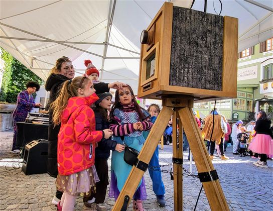 A group of children stands around an old camera and looks curiously. In the background, more people and a festive atmosphere can be seen.