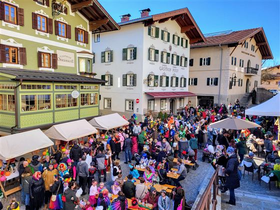 A lively festival with many people in colorful costumes in a square. Surrounded by traditional buildings and cozy seating areas.