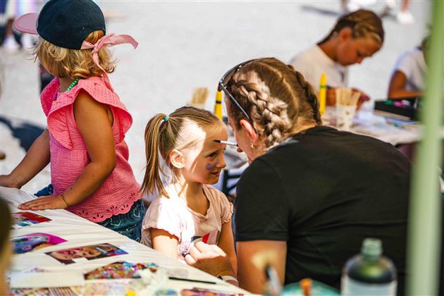 Ein Kind bekommt ein Gesichtsmalerei von einer Betreuerin. Im Hintergrund sind weitere Kinder beim Malen zu sehen.