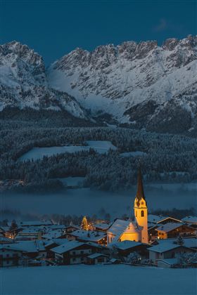 Eine idyllische Berglandschaft mit schneebedeckten Gipfeln und einem kleinen Dorf. Die Kirche im Dorf leuchtet sanft im Dunkeln.