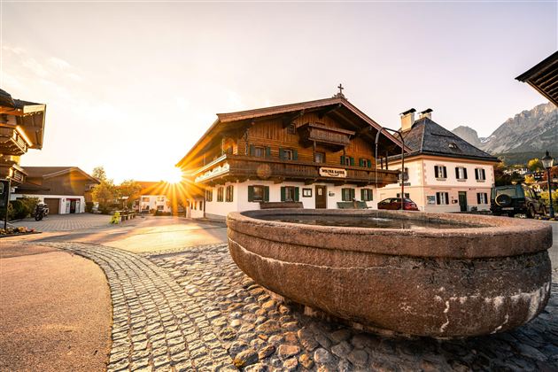 A picturesque landscape with traditional wooden houses and an old well. The sun rises behind the mountains, creating a warm atmosphere.