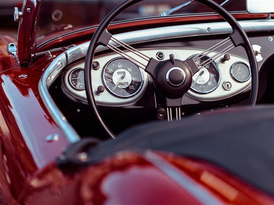 Obrázek zobrazuje interiér klasického červeného sportovního auta s elegantními přístroji. Pohled je zaměřen na volant a palubní desky.