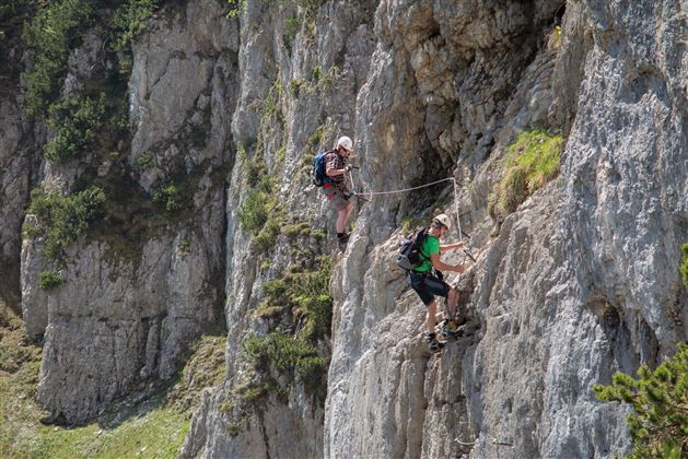 Two climbers rappelling down a rock face. The surroundings are green and mountainous, and the sun is shining.
