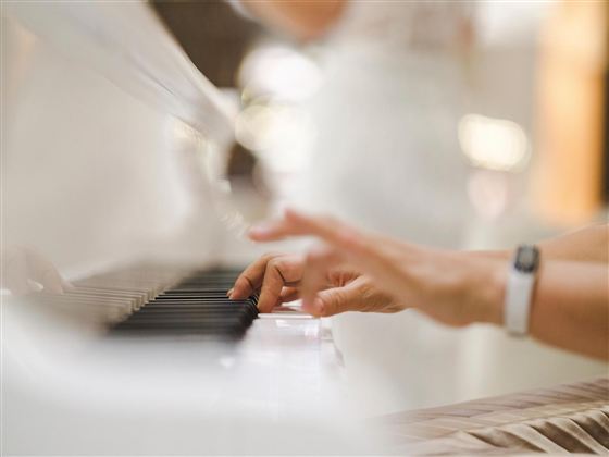 A hand is playing on the keys of a white piano. In the background, a blurry person can be seen.