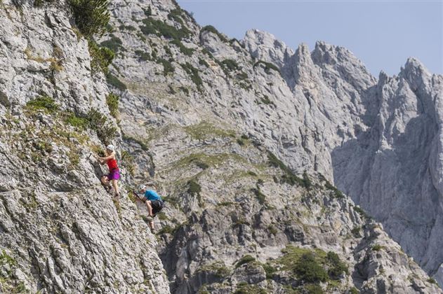 Two climbers are ascending a steep rock wall in an impressive mountain landscape. The sky is clear and the surroundings look majestic.