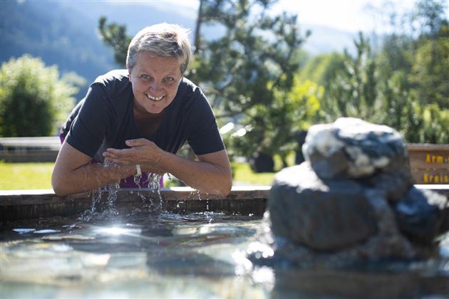Eine Frau lehnt sich über einen kleinen Wasserfall und lächelt in die Kamera. Im Hintergrund sind grüne Bäume und eine hügelige Landschaft zu sehen.