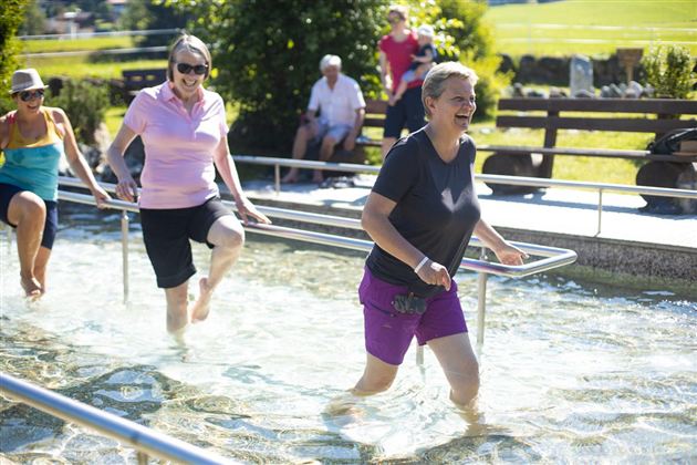 Eine Gruppe von Frauen läuft im Wasser und genießt die warmen Sonnenstrahlen. Im Hintergrund sind Bänke und eine grüne Wiese zu sehen.