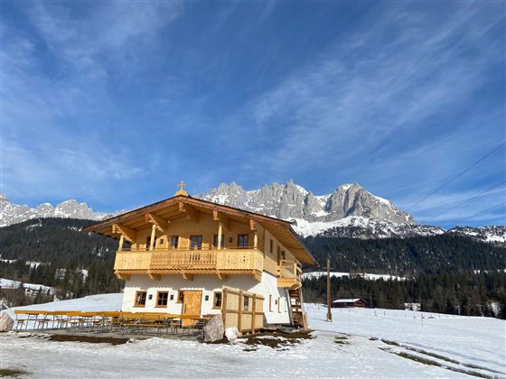 Ein charmantes Holzhaus in den Bergen, umgeben von Schnee. Der klare Himmel und die majestätischen Felsen im Hintergrund schaffen eine beeindruckende Winterlandschaft.