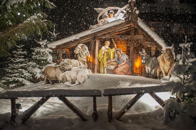 A festive nativity scene in the snow with figures of Mary, Joseph, and the baby Jesus. All around are animals and snow-covered trees.