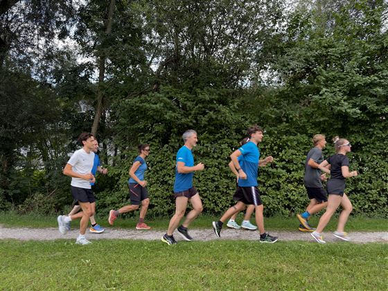 Eine Gruppe von Menschen joggt auf einem schmalen Weg. Umgeben von viel Grün und Bäumen bewegen sie sich in einem aktiven Tempo.