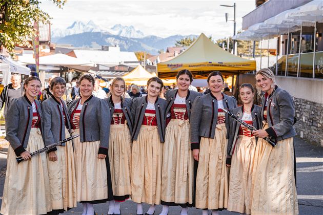 Eine Gruppe von Mädchen und Frauen in traditionellen Trachten steht lächelnd zusammen. Im Hintergrund sind Berge und ein Festzelt zu sehen.